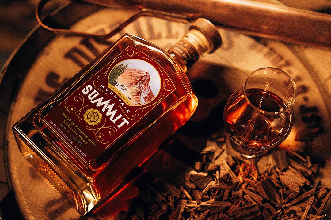 Mountain Summit: Ambruana Finished Whiskey Bottle - Professionally photographed. Sitting atop an aging barrel with a glass next to it.